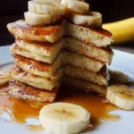 Fluffy homemade banana pancakes stacked on a plate with syrup and bananas